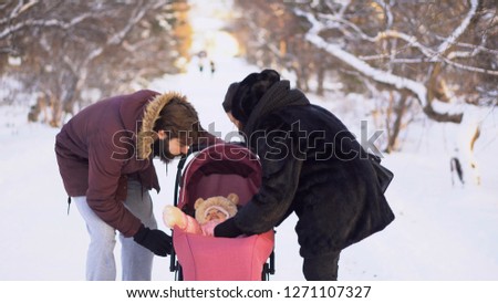 Young couple walking with baby in stroller in winter. Young married couple looks into stroller to happy baby while walking. Happy young family on walk in park in winter