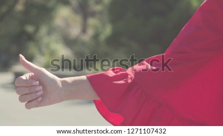 Close-up finger sign up hitchhiking. Stock. Beautiful female hand rises to show sign of hitchhiking. Close-up of woman's hand in red dress against clear blue sky on sunny day