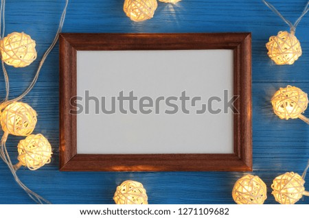 Brown wooden frame with a white paper sheet and cream color fairy lights lit on a blue wooden background. Top view. Flat lay