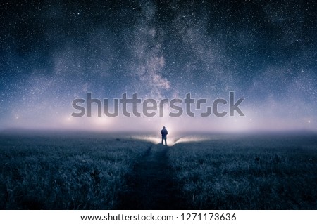 A silhouette of a man with a torch in a field, looking at lights on the horizon on a moody misty night. With a galaxy of stars overhead. Royalty-Free Stock Photo #1271173636