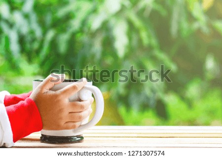 Two hands holding a white mug on wooden table top and green nature background under morning sunlight.