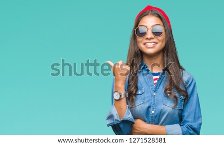 Young beautiful arab woman wearing sunglasses over isolated background smiling with happy face looking and pointing to the side with thumb up.