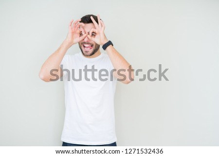Young handsome man wearing casual white t-shirt over isolated background doing ok gesture like binoculars sticking tongue out, eyes looking through fingers. Crazy expression.