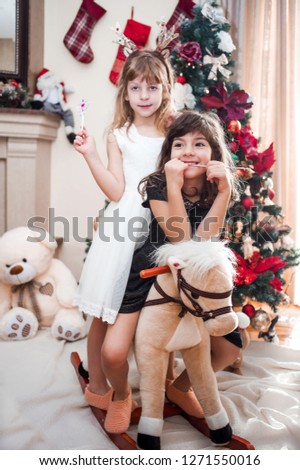 Two adorable, cute girls, sisters, twins in white and black dress with their horse toy, smiling, Xmas tree in the background