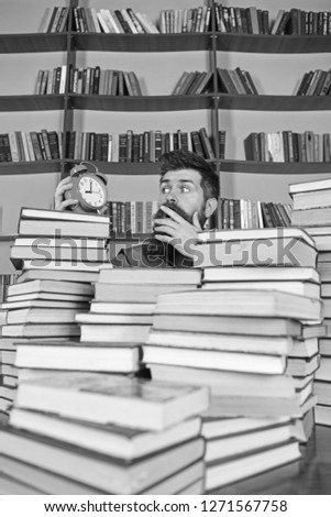 Man, scientist peeking out of piles of books with alarm clock. Teacher or student with beard studying in library. Deadline concept. Man on surprised face looking at clock, bookshelves on background.