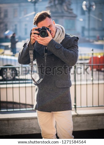 Handsome young male photographer taking photograph with professional photo camera hanging from his neck, outdoor in city street in Europe