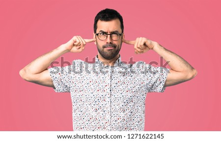 Handsome man with glasses covering both ears with hands on pink background