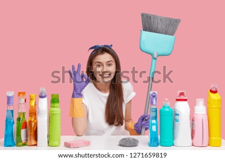 Horizontal view of pleased housemaid makes okay gesture, satisfied with result of perfect cleaning, uses good quality detergents, smiles broadly, isolated over pink background, sits at table