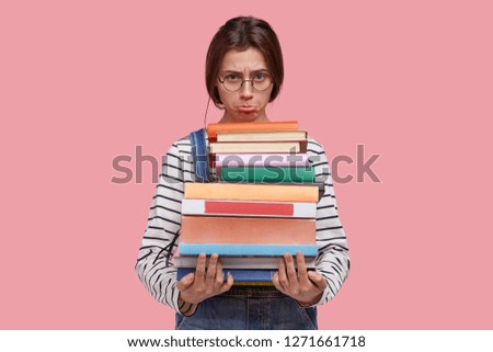 Unhappy sad girl purses lower lip, holds huge pile of books, feels tired of studying and learning much for session, dressed in striped jumper and denim overalls, models against pink background