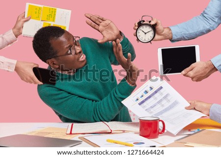 Stressful dissatisfied dark skinned guy makes refusal sign, raises palms over alarm clock, touchpad and papers, has many tasks in one minute, poses at desktop, isolated over pink background. Oh no!