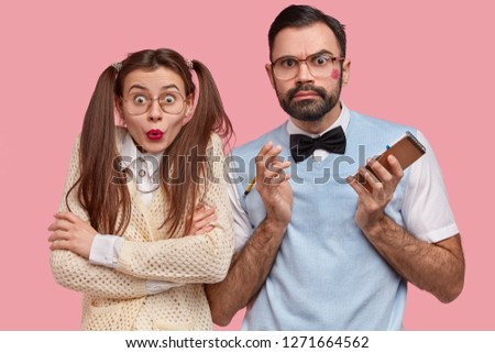 Astonished nerdy couple stare at camera with indignant expressions, recieve bad news, write down notes in spiral notepad, stand closely over pink background. Clever girl with red lipstick, arms folded