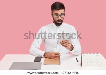 Photo of surprised male employee stares at screen of touchpad, feels astonishment, reads shocking message, drinks coffee from disposable cup, wears spectacles, white shirt, poses alone at desktop