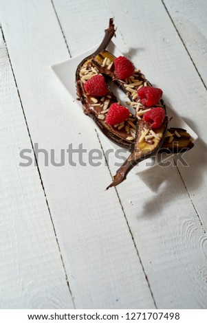 Baked banana dessert with almonds and chocolate with raspberries on old white wooden table.