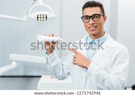 handsome african american doctor in glasses pointing at toothpaste in dental clinic 
