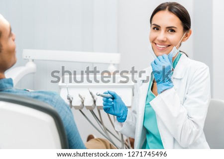 cheerful female dentist holding drill and smiling near patient  Royalty-Free Stock Photo #1271745496