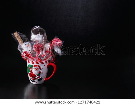 White Christmas mug with Santa Claus picture filled with wrapped cake pops, marshmallows and candy canes on a black background with text space. Christmas baked gifts.