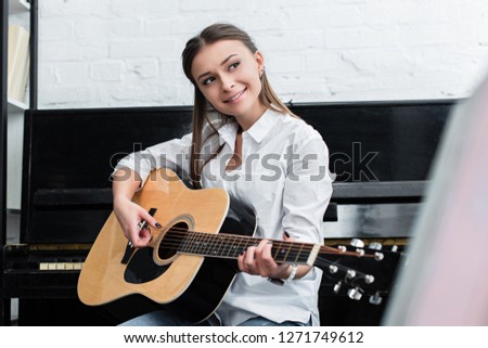 smiling girl sitting and playing guitar in living room with piano on background