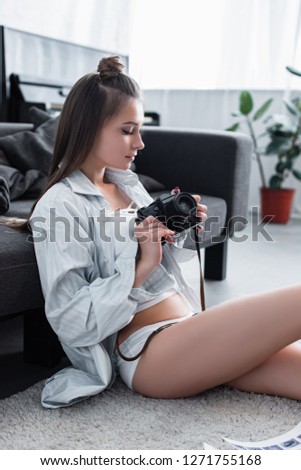 beautiful photographer in shirt and underwear sitting on carpet near sofa and holding vintage film camera in living room
