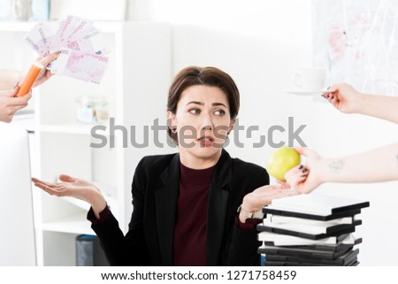 attractive businesswoman gesturing while secretaries holding money, cup and apple in office