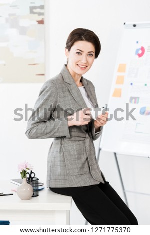 attractive businesswoman in grey suit holding cup of coffee ans sitting on table in office