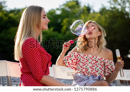 Two attractive beautiful young blond women blow bubbles in the park and are happy on sunset. Girlfriends in park.
