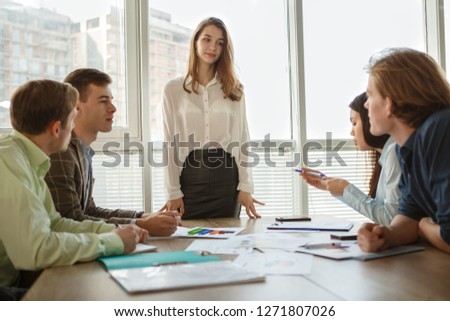 Lady boss standing near table and presenting new project to workers in office. Smart colleagues sitting ans looking at her and listening carefully. Concept of cooperation and teamwork.