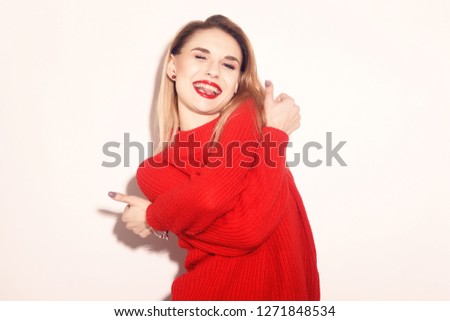 Happy Smiling Girl in knitted red sweater having fun . Cute blond woman Warming Up Concept. Pretty girl with empty copy space near. Beautiful teen girl with red lips. Girl showing tongue and thumbs up