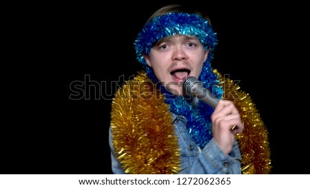 Cheerful, positive, handsome man holding microphone, listening to music and singing song with tinsel, isolated on black background. Funny man in karaoke club singing with tinsel
