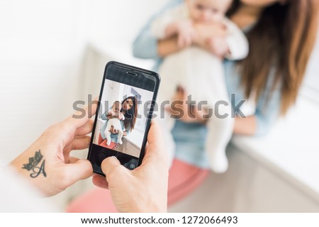 cropped shot of man taking picture of wife with baby in hands on smartphone