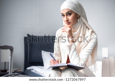 young muslim woman sitting on bed and looking at camera while writing in notebook