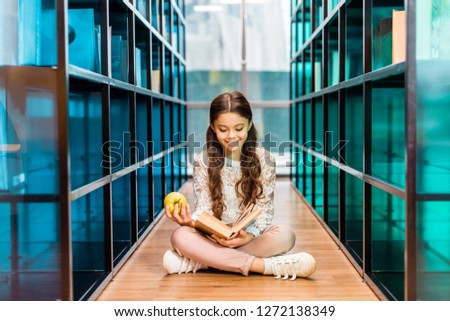 Beautiful happy schoolchild holding apple and reading book in library