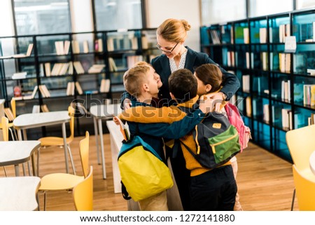 happy schoolchildren with backpacks hugging librarian in library 