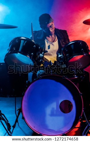 handsome male musician in leather jacket playing drums during rock concert on stage with smoke and dramatic lighting