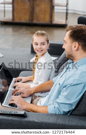 smiling father and daughter sitting on sofa, typing on laptop keyboards and looking at each other