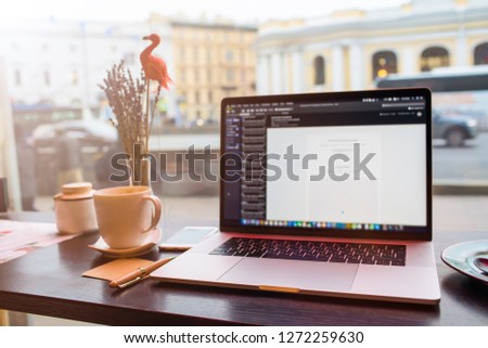Close up of open modern pc laptop computer with blurred applications on the screen standing on wooden table near cup of coffee against restaurant window with street view. Notebook device 