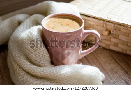 A Cup of hot cappuccino coffee with wicker basket and warm scarf a on wooden table.  Winter mood concept. Autumn mood concept. Warm autumn or winter picture. Selective focus. At home. Time for relax.