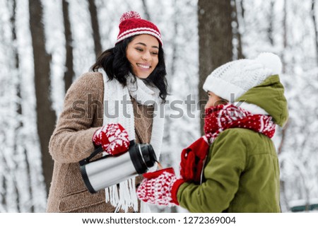 african american mother smiling and pouring tea from thermos in red cup holding by daughter in winter park