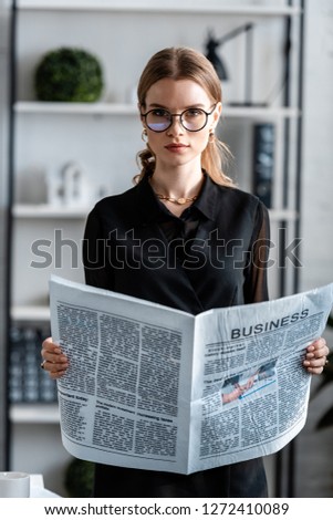 attractive businesswoman in black clothes and glasses holding newspaper and looking at camera