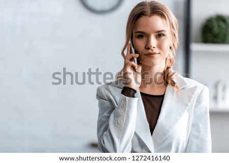 businesswoman in white formal wear talking on smartphone and looking at camera at workplace