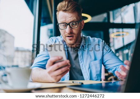 Excited young man blogger reading shocked news on internet website on modern smartphone device using free 4G internet connection.Amazed hipster guy watching video in social networks on cellular