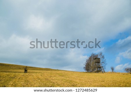 Spring landscape with brown field, trees and wooden high seat on horizon, blue and white cloudy sky, copy space