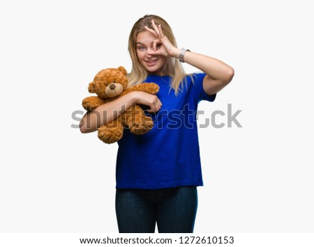 Young caucasian woman holding cute teddy bear over isolated background with happy face smiling doing ok sign with hand on eye looking through fingers