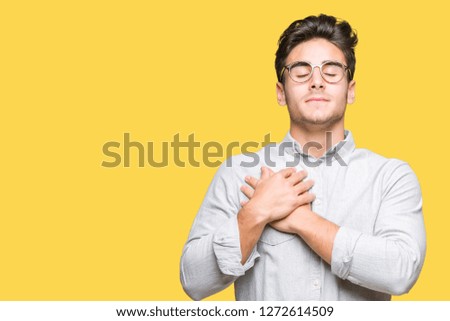 Young handsome man wearing glasses over isolated background smiling with hands on chest with closed eyes and grateful gesture on face. Health concept.