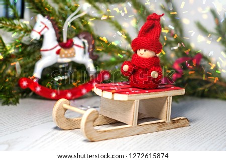 Christmas card. Bright red toy kid in knitted clothes on Santa's sleigh on the background of a Christmas tree and a toy horse.