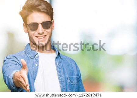 Young handsome man wearing sunglasses over isolated background smiling friendly offering handshake as greeting and welcoming. Successful business.