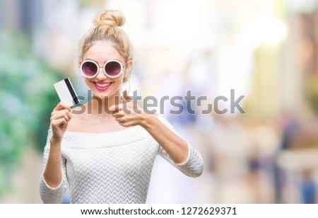 Young beautiful blonde woman holding credit card over isolated background happy with big smile doing ok sign, thumb up with fingers, excellent sign