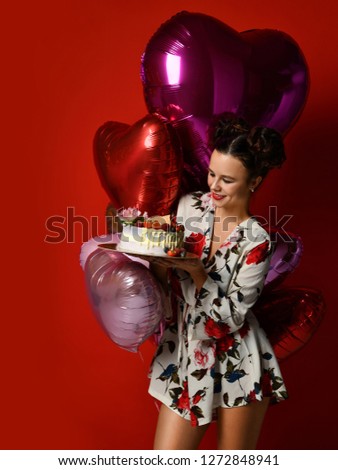 Young woman hold big sweet cake with strawberry blueberry and cream smiling celebrating birthday with balloons on dark red background 