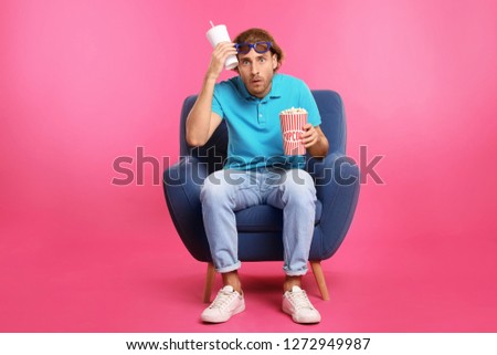 Emotional man with 3D glasses, popcorn and beverage sitting in armchair during cinema show on color background