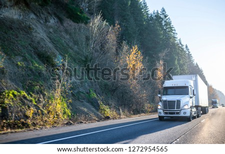 Big rig bright white classic semi truck with day cab for local haul routs transporting dry van semi trailer driving on the straight highway in front of another traffic in sunny day Royalty-Free Stock Photo #1272954565
