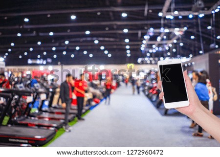 Hand holding mobile phone with abstract blurred background image and bokeh light of crowd people at cars exhibition show, internet and social network concept